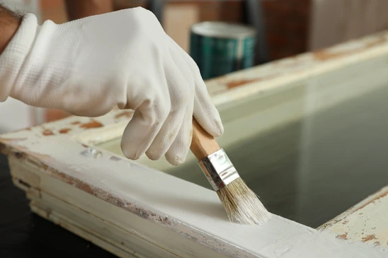 Intérieur d'atelier, personne portant gants blancs appliquant peinture au pinceau sur meuble en bois clair