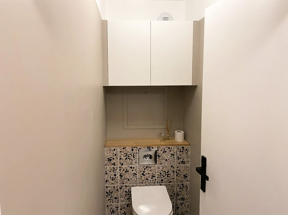 WC suspendu avec façade en carreaux de terrazzo noir et beige, plateau en bois chêne naturel, bouton de chasse chromé encastré