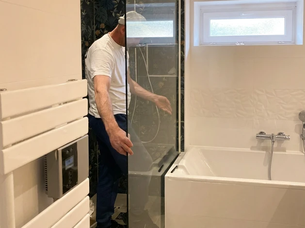 Un homme en polo blanc et jean gris travaille à genoux sur l'installation d'une porte intérieure dans un appartement moderne aux murs blancs