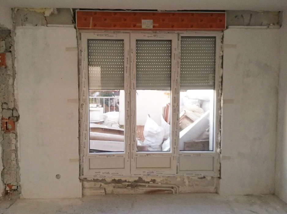 Un homme en polo blanc et jean gris travaille à genoux sur l'installation d'une porte intérieure dans un appartement moderne aux murs blancs